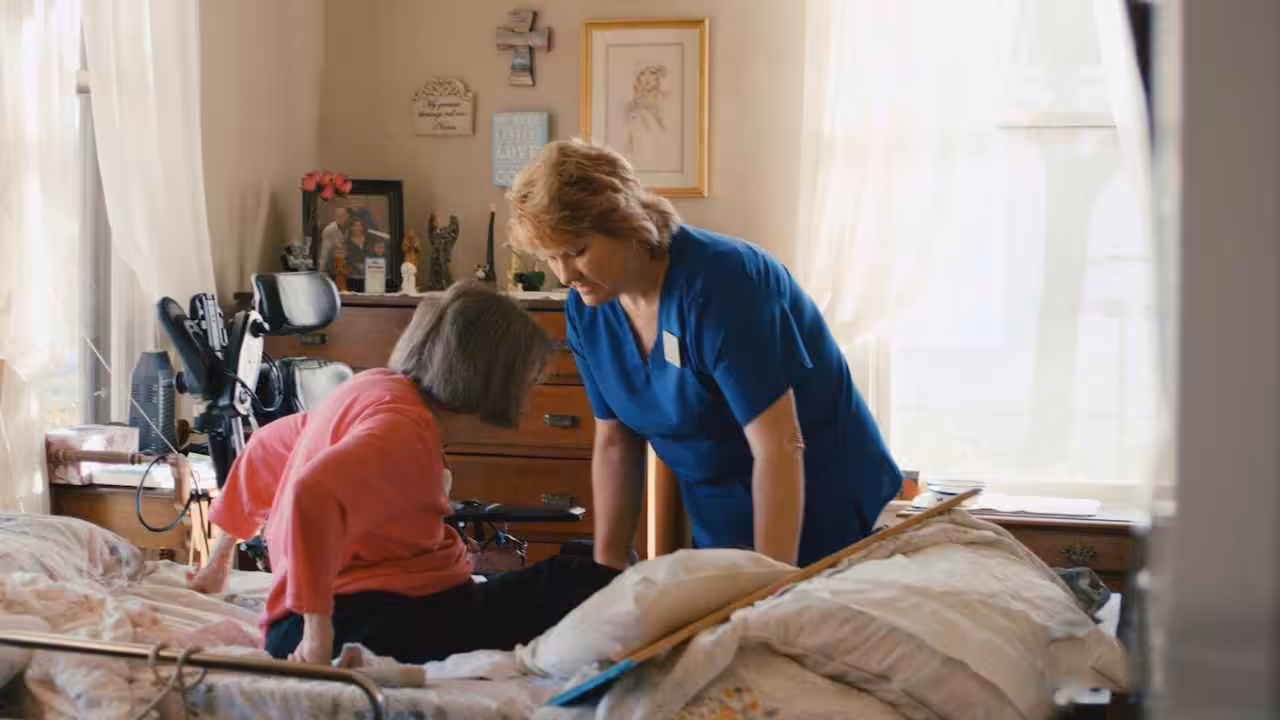 A caregiver in blue scrubs assists an elderly woman sitting on a bed in a cozy, sunlit bedroom with medical equipment and family photos visible in the background.