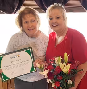 Two women smiling indoors; one holds a bouquet of red and white flowers, the other holds a framed certificate that reads “Exceptional Caregiver Nominee.” They stand close together in front of a bright window.