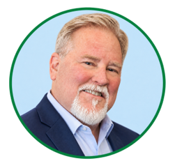 Smiling older man with light hair, short beard, and mustache, wearing a navy blazer and light blue shirt. Light blue background with a green circular border.