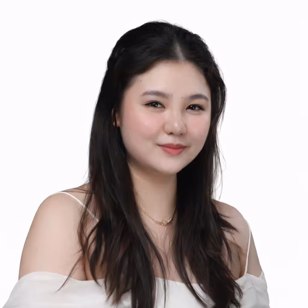 A young woman with long dark hair and fair skin smiles gently. She is wearing an off-shoulder white top, a delicate necklace, and has a natural makeup look. The background is plain white.