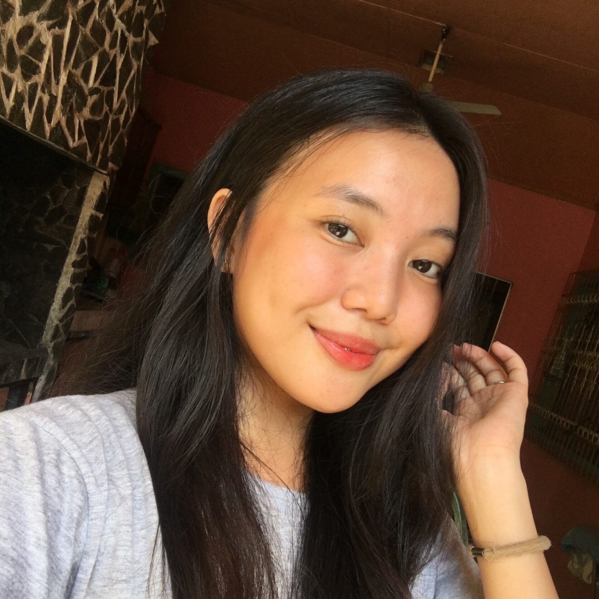 A young woman with long dark hair and a gray shirt smiles while taking a selfie indoors, with sunlight on her face and a stone wall in the background.