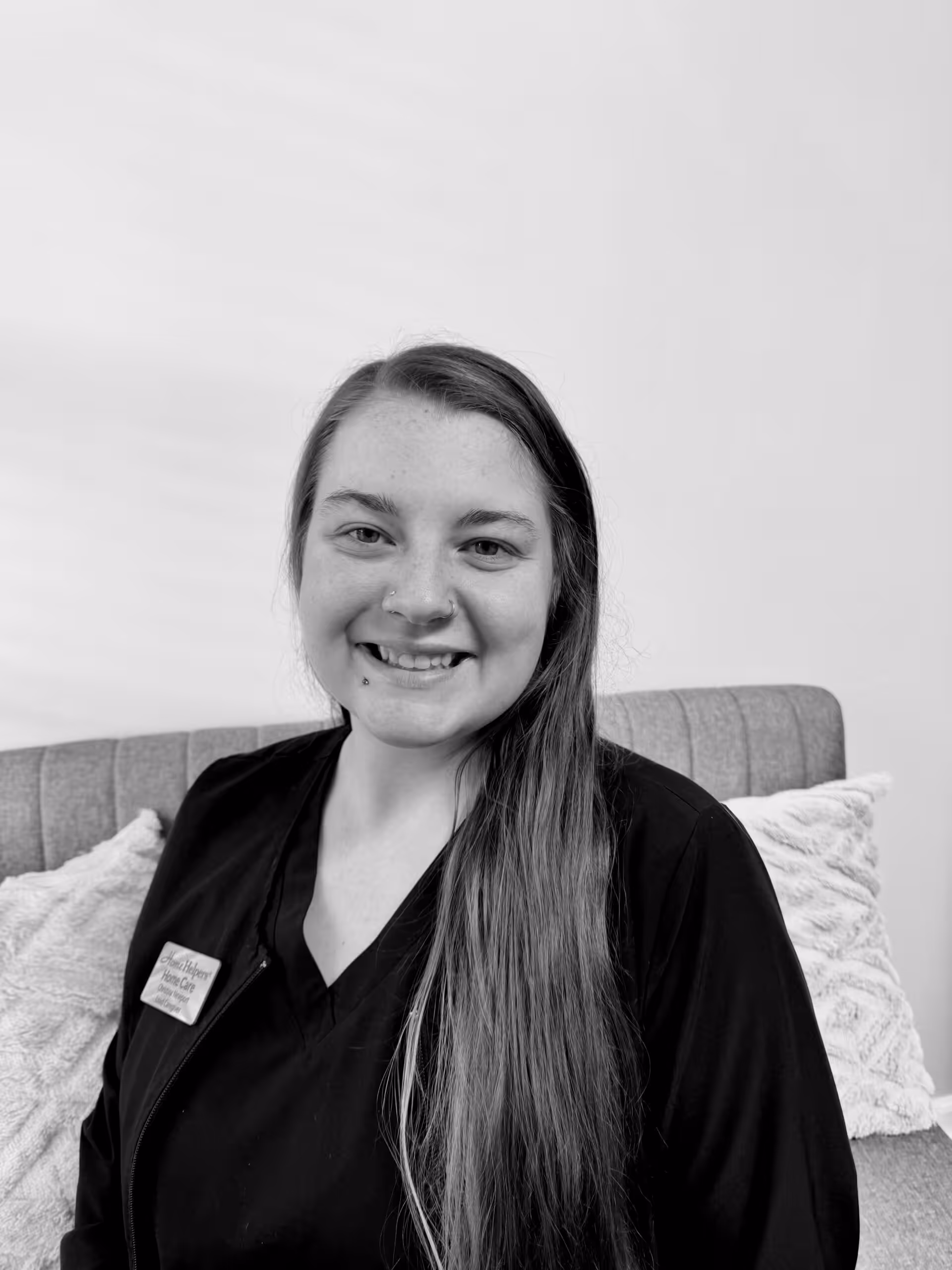 A woman with long hair wearing a dark top and a name tag sits on a cushioned bench, smiling at the camera. Light-colored pillows are behind her, and the background is plain and bright. The photo is in black and white.