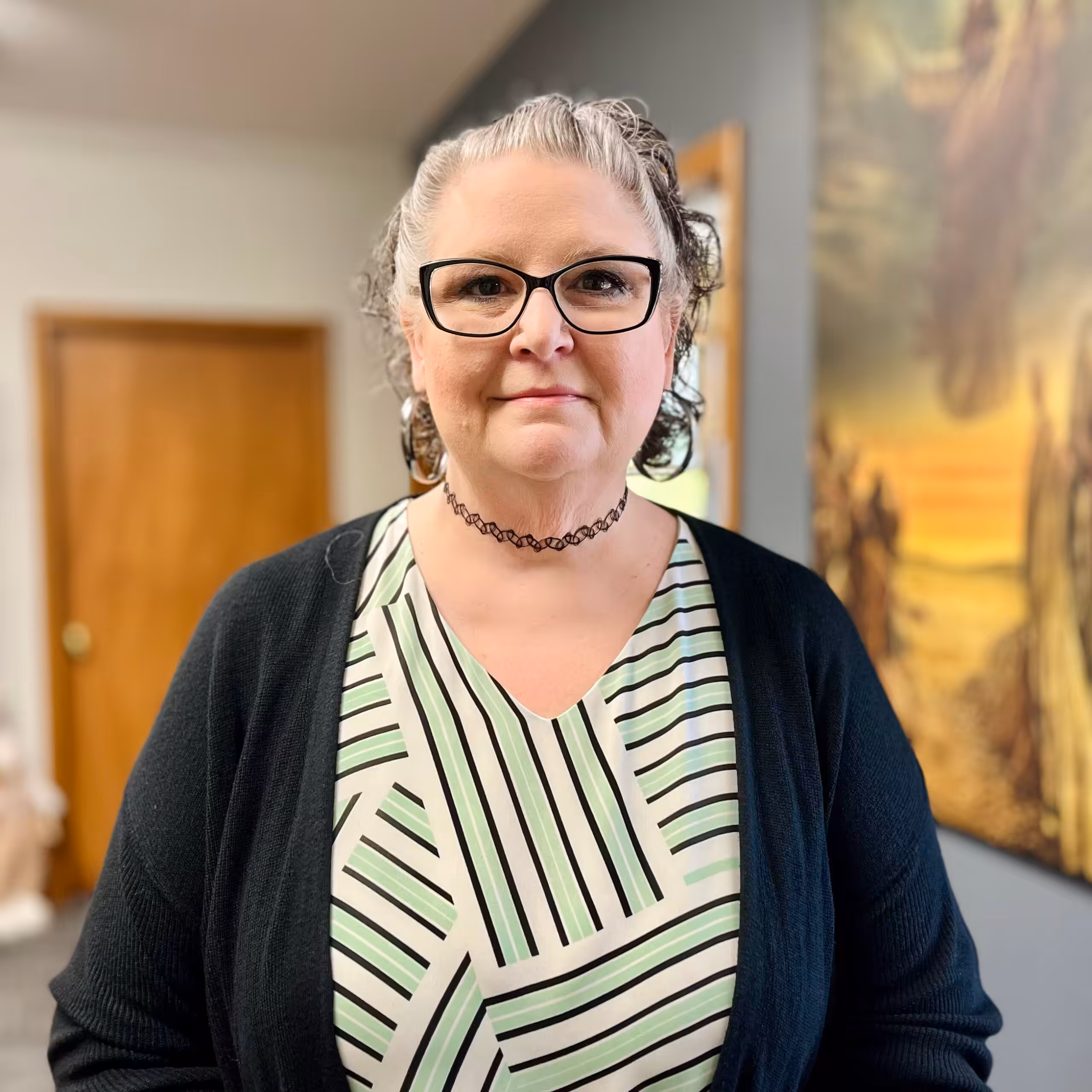 A woman with short, curly gray hair and glasses stands indoors, wearing a black cardigan and a striped dress with a black choker necklace. A wooden door and a painting are visible in the background.