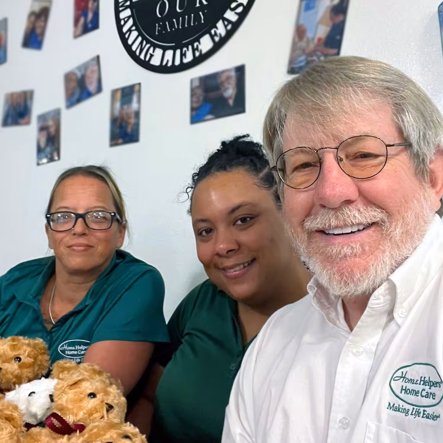 Three smiling adults, two women in green shirts and a man in glasses and a white shirt, sit together holding teddy bears. Photographs and a sign are displayed on the wall behind them.