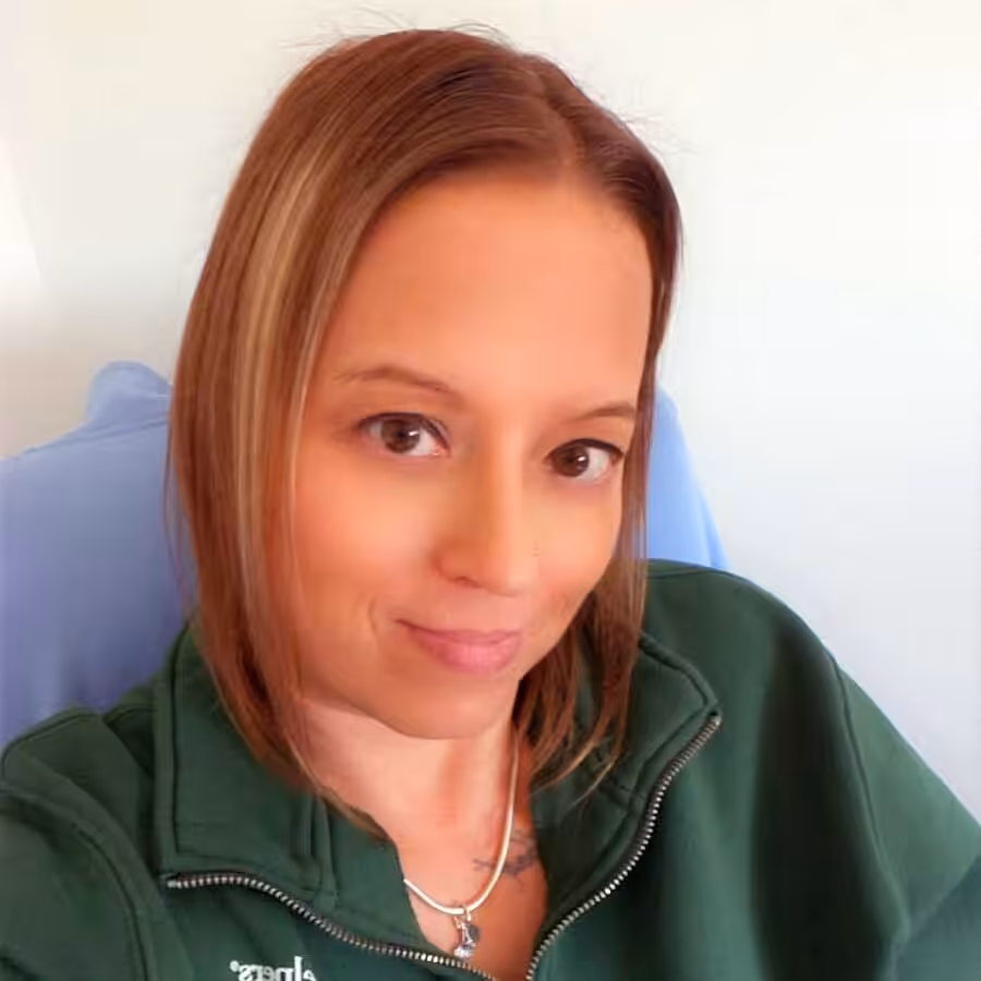 A woman with straight, shoulder-length light brown hair is sitting against a light background, wearing a green zip-up top and a necklace. She is looking at the camera with a slight smile.
