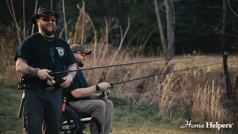 Two men, one standing and one in a wheelchair, smile while fishing outdoors near tall grass and trees. Both wear hats and sunglasses. The Home Helpers logo is in the bottom right corner.