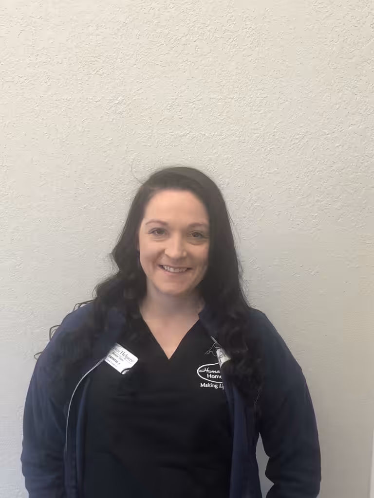 A woman with long, dark wavy hair wearing a dark jacket and black shirt stands smiling in front of a plain light-colored wall. She has a name tag and a logo on her shirt.