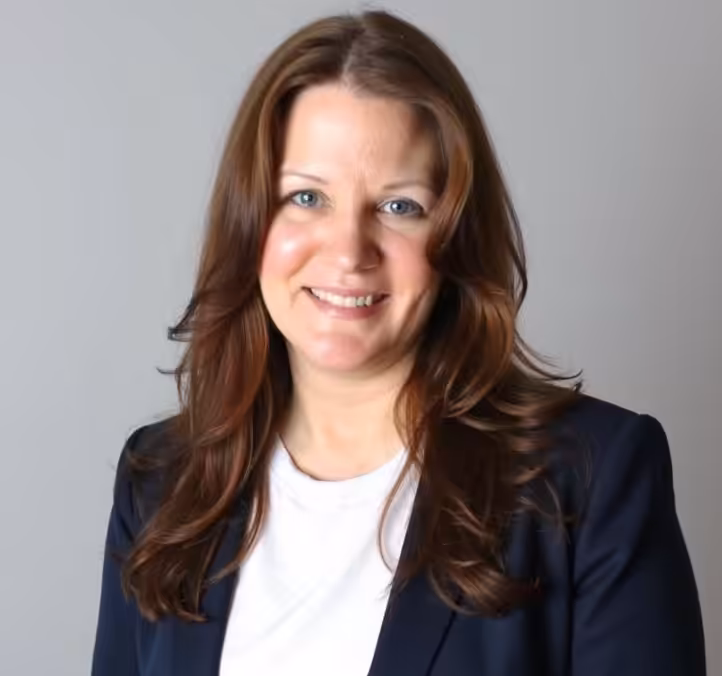 A woman with long brown hair wearing a navy blazer over a white top smiles at the camera against a plain gray background.