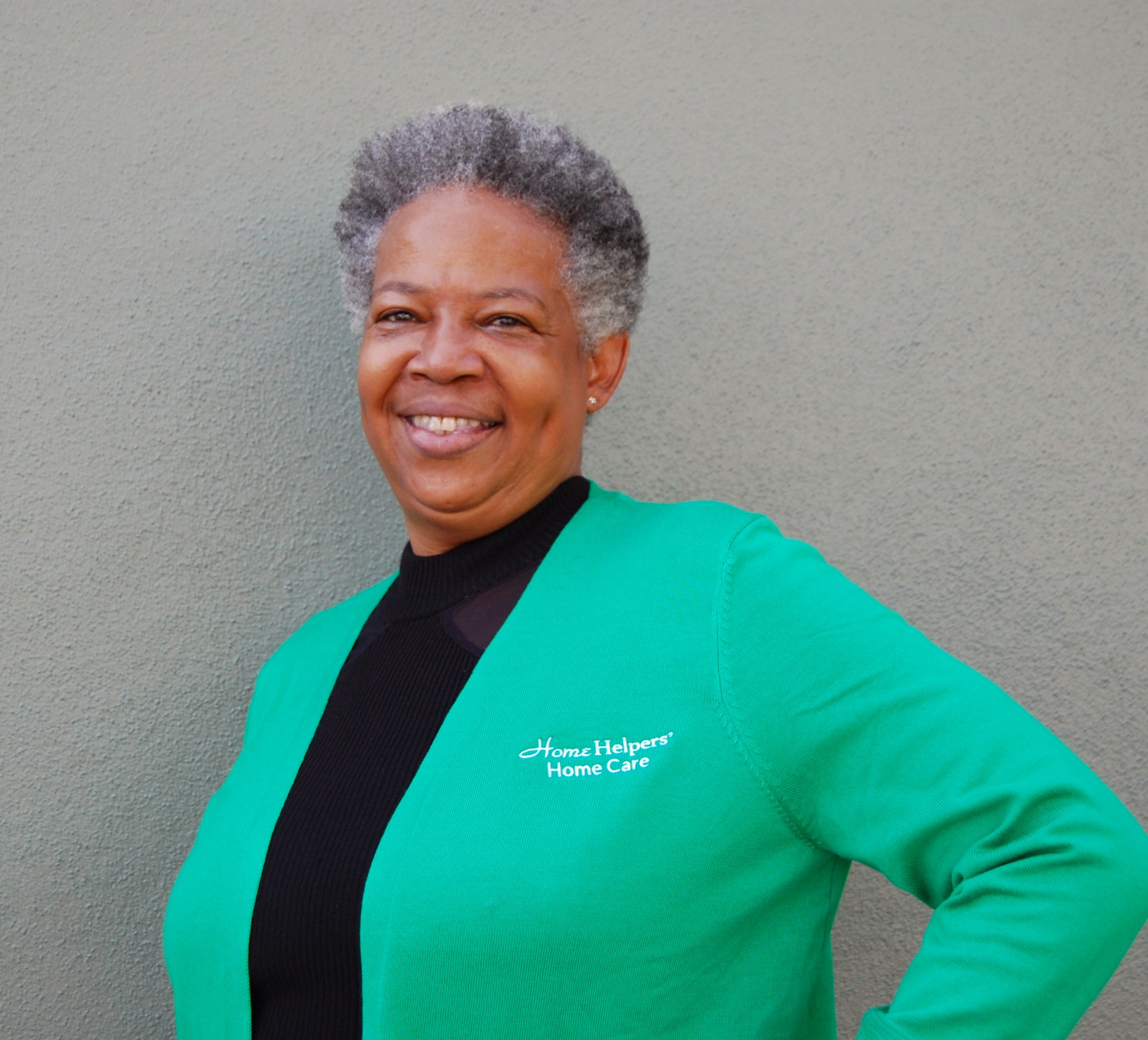 Smiling person with short gray hair wearing a green Home Helpers Home Care sweater, standing against a plain gray wall with one hand on their hip.