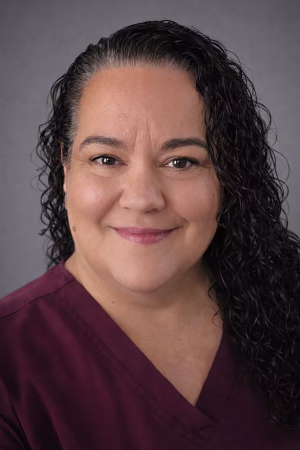 A woman with long, curly dark hair smiles gently at the camera. She is wearing a maroon scrub top and is posed in front of a plain gray background.