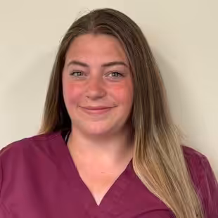 A woman with long, straight, light brown hair wearing a maroon scrub top stands in front of a plain light-colored wall, smiling gently at the camera.