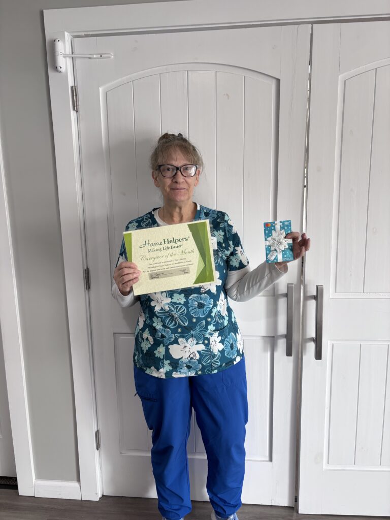 A woman in blue scrubs with a floral top stands in front of white double doors, holding a certificate and a gift card, smiling at the camera.