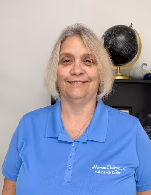 A woman with short blonde hair wearing a blue Home Helpers polo shirt stands indoors, smiling. A shelf with a black globe and decorative items is visible in the background.