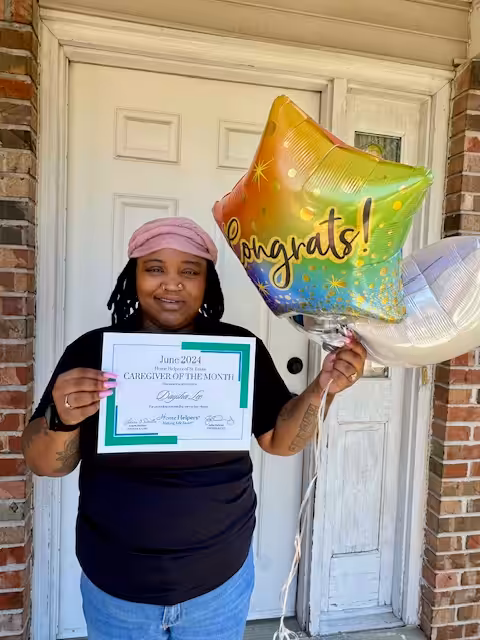 A smiling person stands in front of a door, holding a Congrats! balloon and a certificate that reads June 2024 Caregiver of the Month. They are wearing a pink headscarf and a black shirt.
