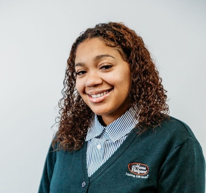 A woman with curly brown hair smiles, wearing a striped shirt and a dark green Home Helpers sweater against a plain light background.