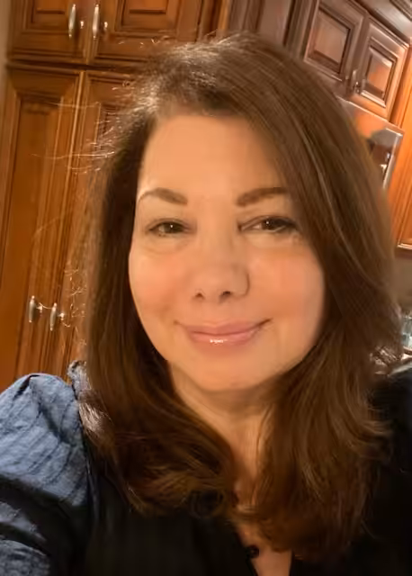 A woman with long brown hair and a light pink lipstick smiles softly while taking a selfie in a kitchen with wooden cabinets in the background.
