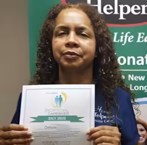 A woman with curly hair holds a certificate and stands in front of a green and white Helper-branded banner. She is wearing a blue shirt and looking at the camera.