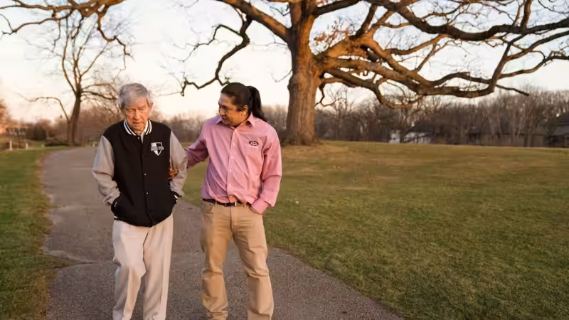 Two men walk together on a paved path in a park. One man, older and wearing a black jacket, is being gently supported by the other, who wears a pink shirt. A large leafless tree stands in the background.