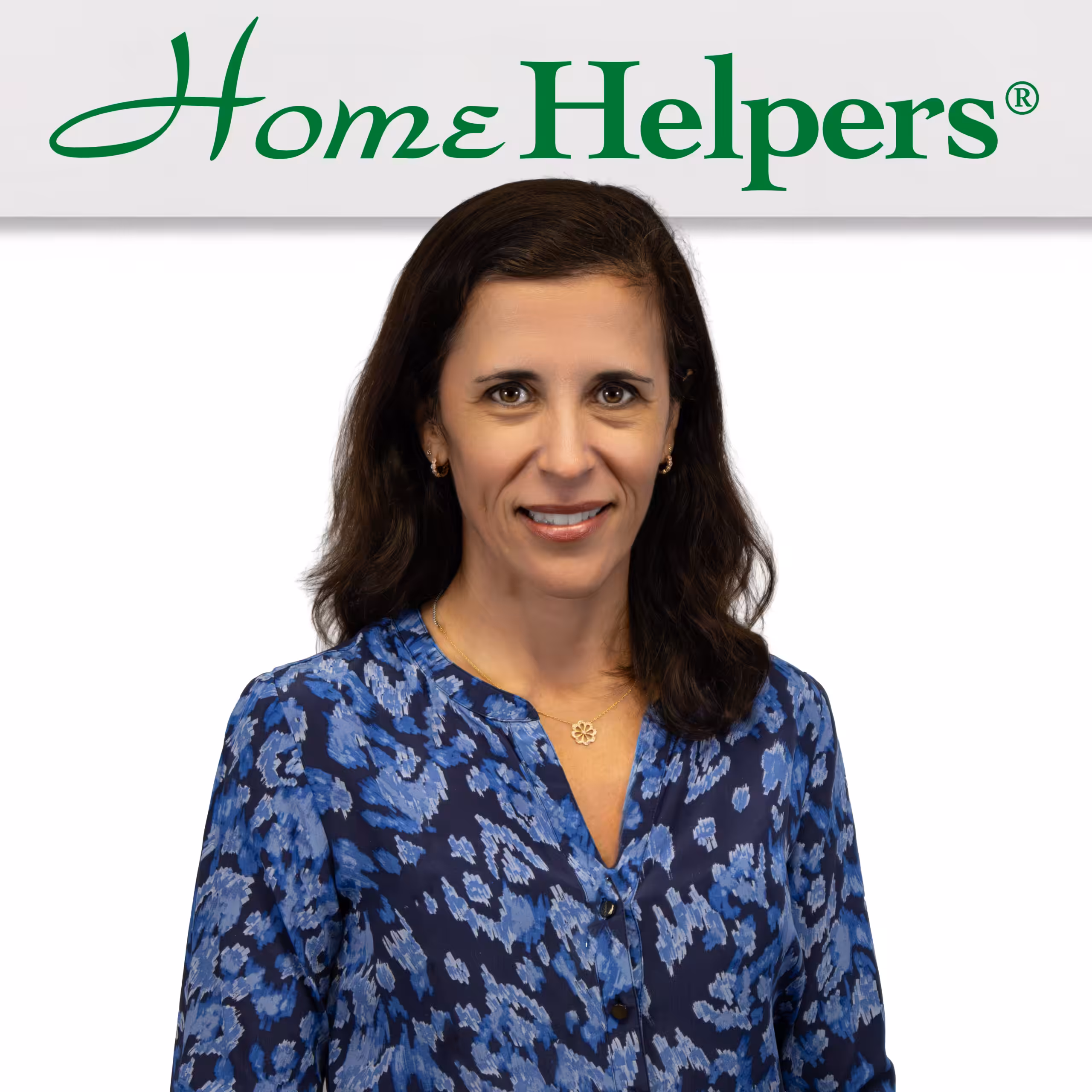 A woman with long dark hair, wearing a blue patterned blouse, stands smiling in front of a white background with the green Home Helpers logo above her.