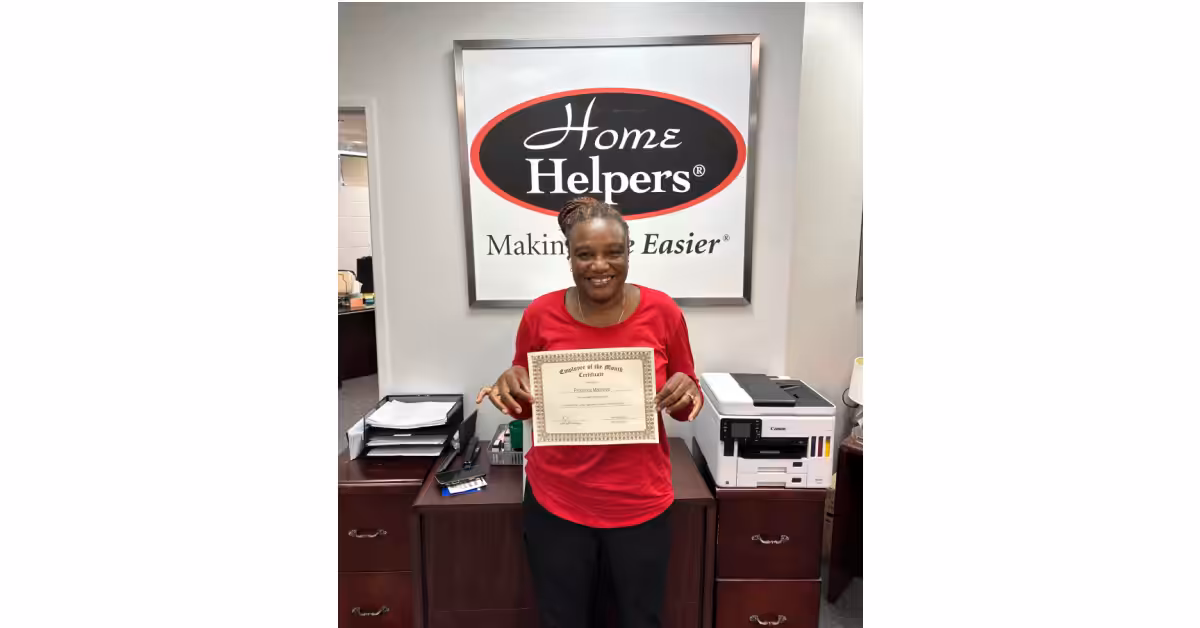 A woman in a red shirt stands in an office, smiling and holding a certificate. Behind her is a sign that reads “Home Helpers Making Life Easier.” Office furniture and a printer are visible in the background.