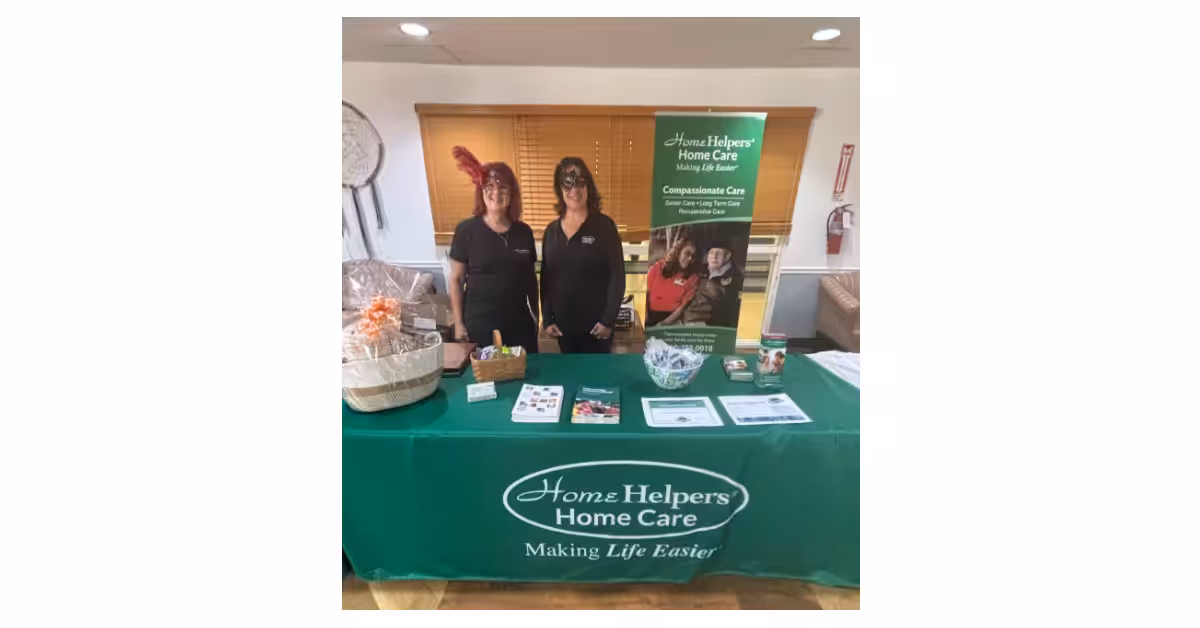 Two women stand behind a table covered with a green Home Helpers Home Care tablecloth, displaying brochures, a gift basket, and promotional materials at an indoor event. A large banner is behind them.
