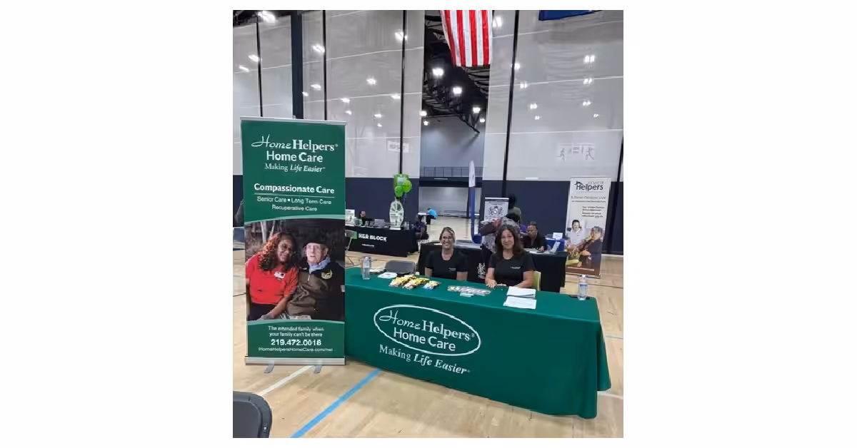 Three people sit behind a green Home Helpers Home Care table at an indoor event. The table has pamphlets and snacks, with a banner and informational materials displayed. An American flag hangs above in the gymnasium.