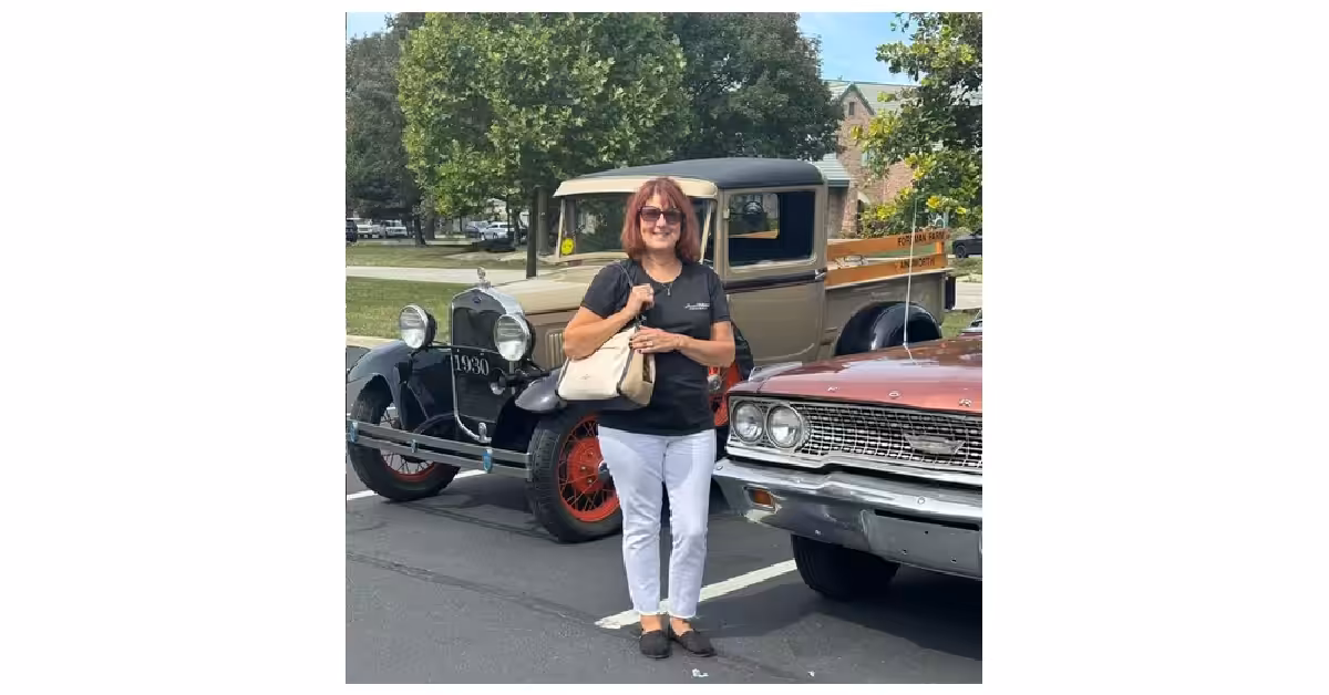 A woman with shoulder-length hair, sunglasses, and a black shirt stands smiling between two vintage cars in a parking lot, holding a beige purse. Trees and a building are visible in the background.