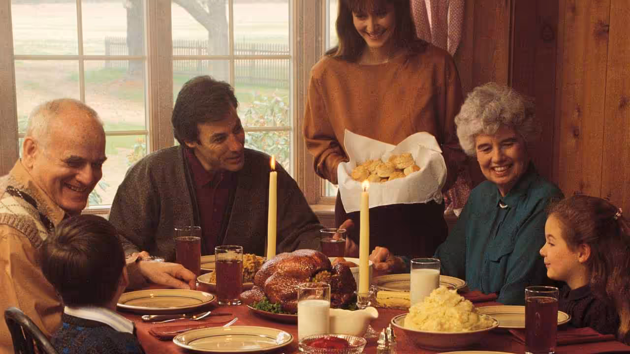 A family gathers around a dinner table set with a turkey, mashed potatoes, and candles. Six people, including children and seniors, smile and talk as a woman serves bread rolls. The scene is warm and cozy inside a wood-paneled room.