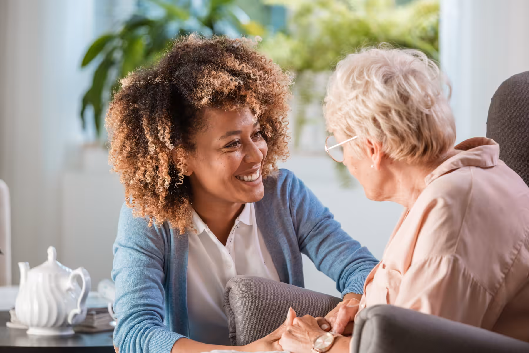 A younger woman smiles warmly and holds hands with an older woman, both seated indoors near a window with plants. A teapot and cup are on a table beside them, suggesting a caring and supportive conversation.