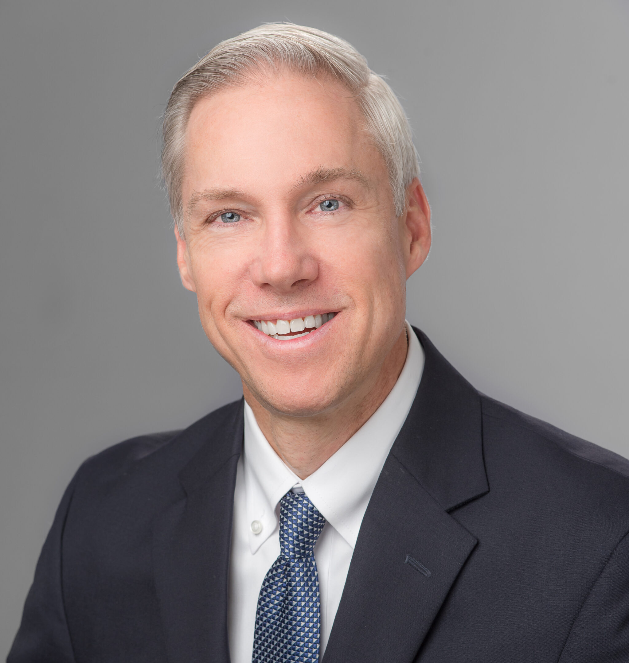 A middle-aged man with short, gray hair wearing a dark suit, white shirt, and patterned tie, smiling in a professional headshot against a plain gray background.