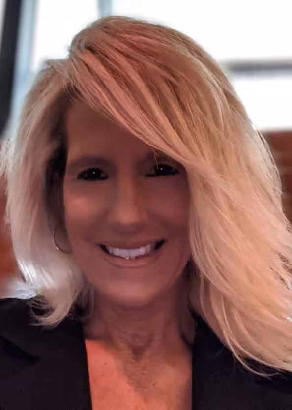 A woman with shoulder-length blonde hair smiles at the camera. She is wearing a dark top and hoop earrings. The background is softly blurred, drawing focus to her face.