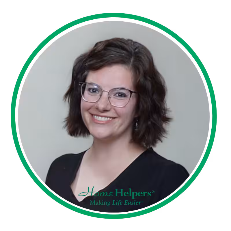 A woman with short brown wavy hair and glasses smiles at the camera. She is wearing a black top. The image is framed by a green double circle with the Home Helpers® Making Life Easier® logo at the bottom.