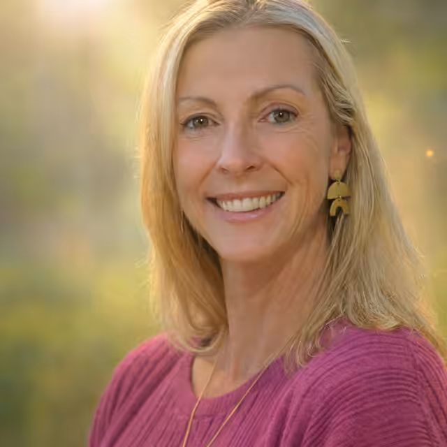 A smiling woman with long blonde hair, wearing a mauve sweater and gold earrings, stands outdoors in soft sunlight with a blurred green background.
