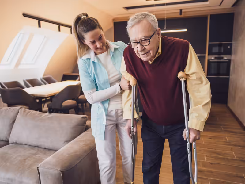 A smiling young woman helps an elderly man using crutches walk through a modern living room. The man appears to be concentrating while the woman offers support by holding his arm.