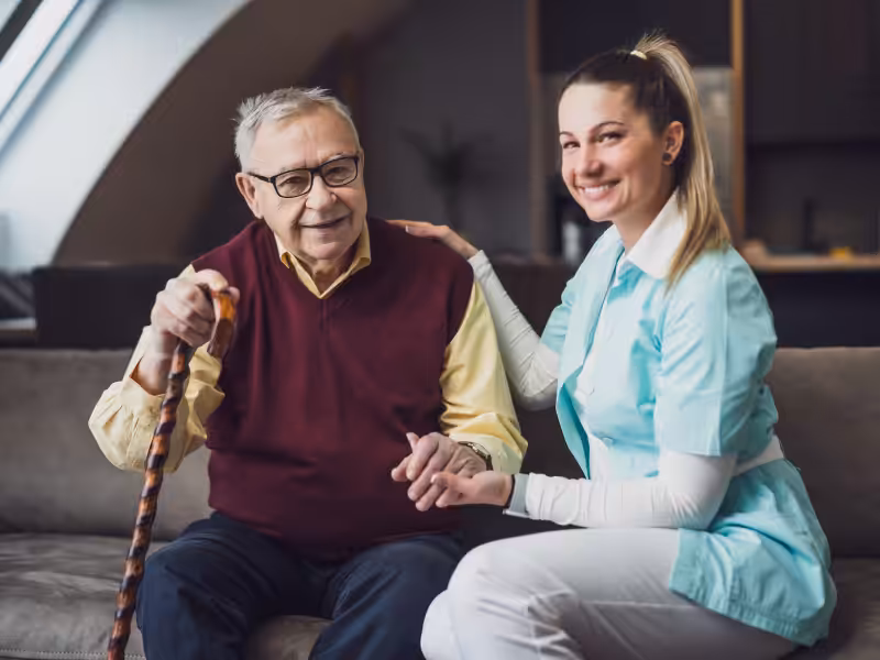 An elderly man with glasses and a cane sits on a sofa, smiling, while a female caregiver in a blue uniform kneels beside him, holding his hand and smiling at the camera.