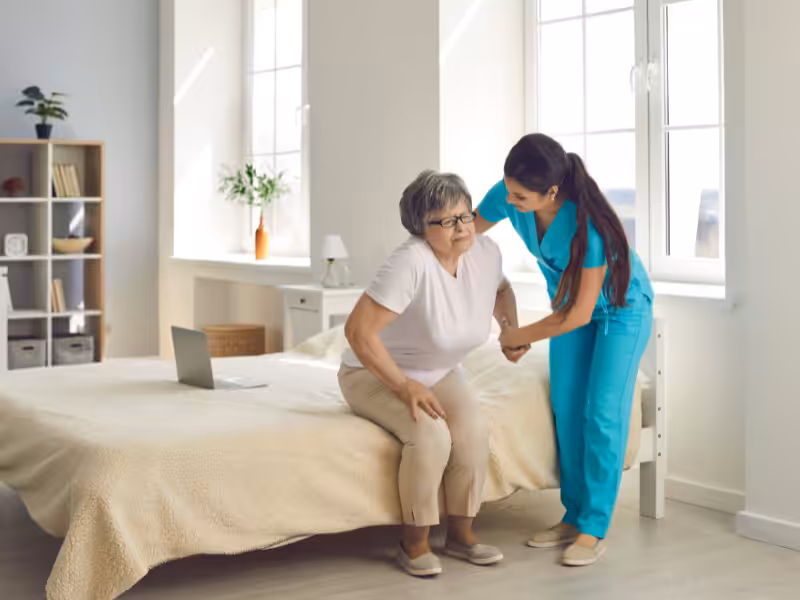 A nurse in blue scrubs helps an older woman stand up from a bed in a bright, tidy bedroom. The woman looks uncomfortable, and the nurse supports her gently by the arm.