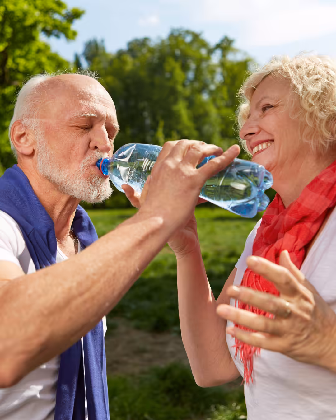 An older woman smiles as she helps an older man drink from a water bottle outdoors on a sunny day, with green trees and grass in the background.
