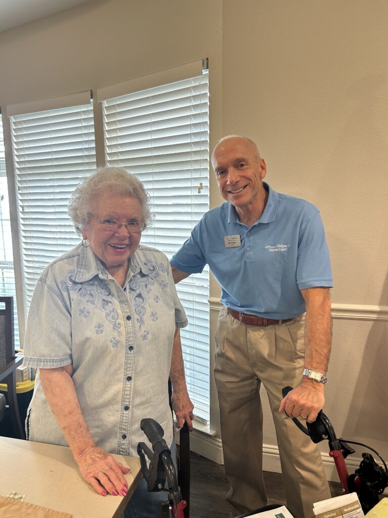 An older woman and a man stand smiling indoors next to each other, each holding a walker. The woman wears a light denim shirt and glasses; the man wears a light blue polo and khaki pants. They are in a well-lit room with shuttered windows.