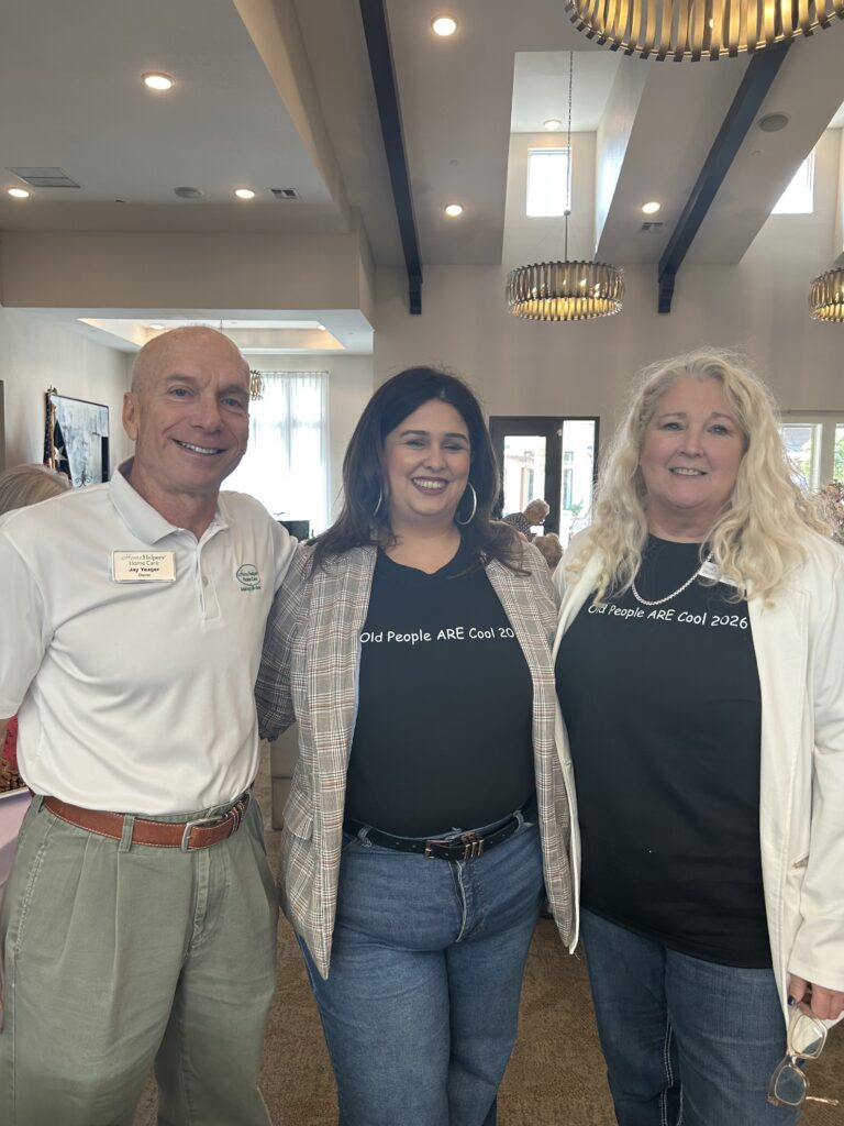 Three adults stand smiling together indoors. Two women wear black shirts reading Old People ARE Cool 2024; the man wears a white polo and name tag. The setting is well-lit with high ceilings and modern chandeliers.