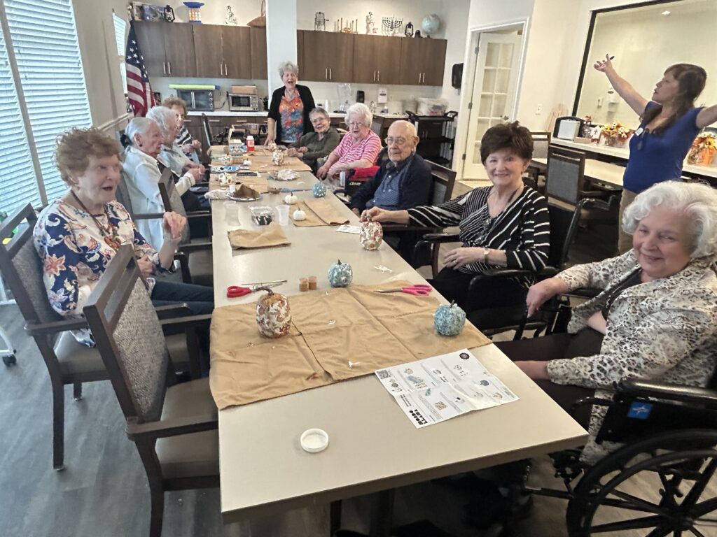 A group of seniors sits around a long table in a bright room, smiling and socializing. Craft supplies and decorative items are on the table. An American flag and two women standing are visible in the background.