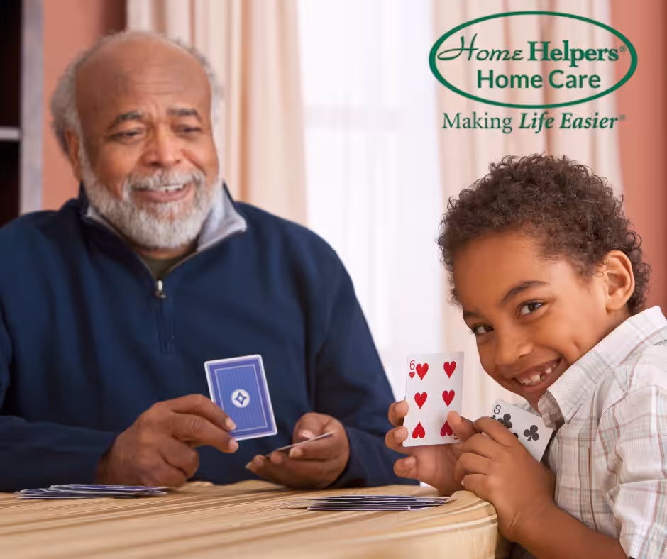 An elderly man and a young boy are smiling and playing cards together at a table. The boy is holding playing cards and looking at the camera. The Home Helpers Home Care logo is in the upper right corner.