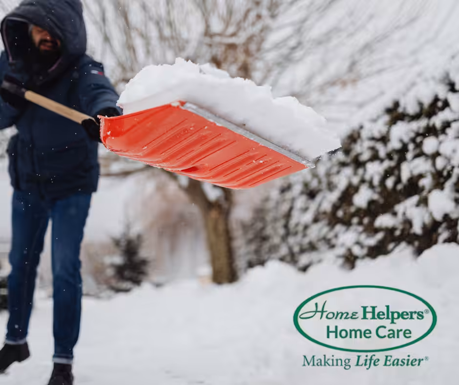 A person in winter clothing shovels snow with a red shovel in a snowy outdoor setting. The Home Helpers Home Care logo and the slogan Making Life Easier appear in the lower right corner.