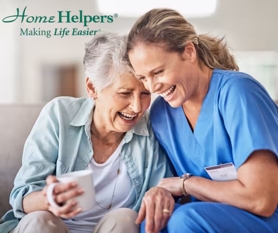 An older woman and a caregiver in blue scrubs sit closely together on a couch, smiling and laughing. The older woman holds a mug. The Home Helpers logo and the phrase Making Life Easier appear in the top left corner.