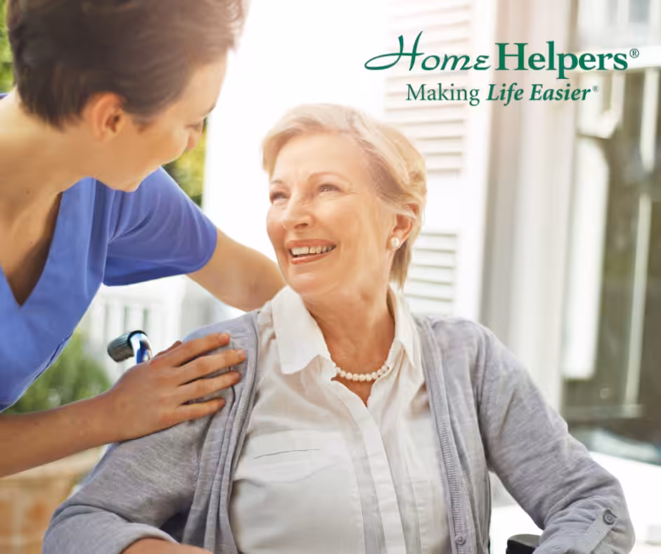 A smiling elderly woman in a wheelchair is comforted by a caregiver in blue scrubs. The caregiver has a hand on her shoulder. The text Home Helpers Making Life Easier is in the upper right corner.