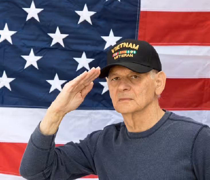 An older man in a Vietnam Veteran cap salutes in front of a large American flag, wearing a dark long-sleeve shirt and a serious expression.