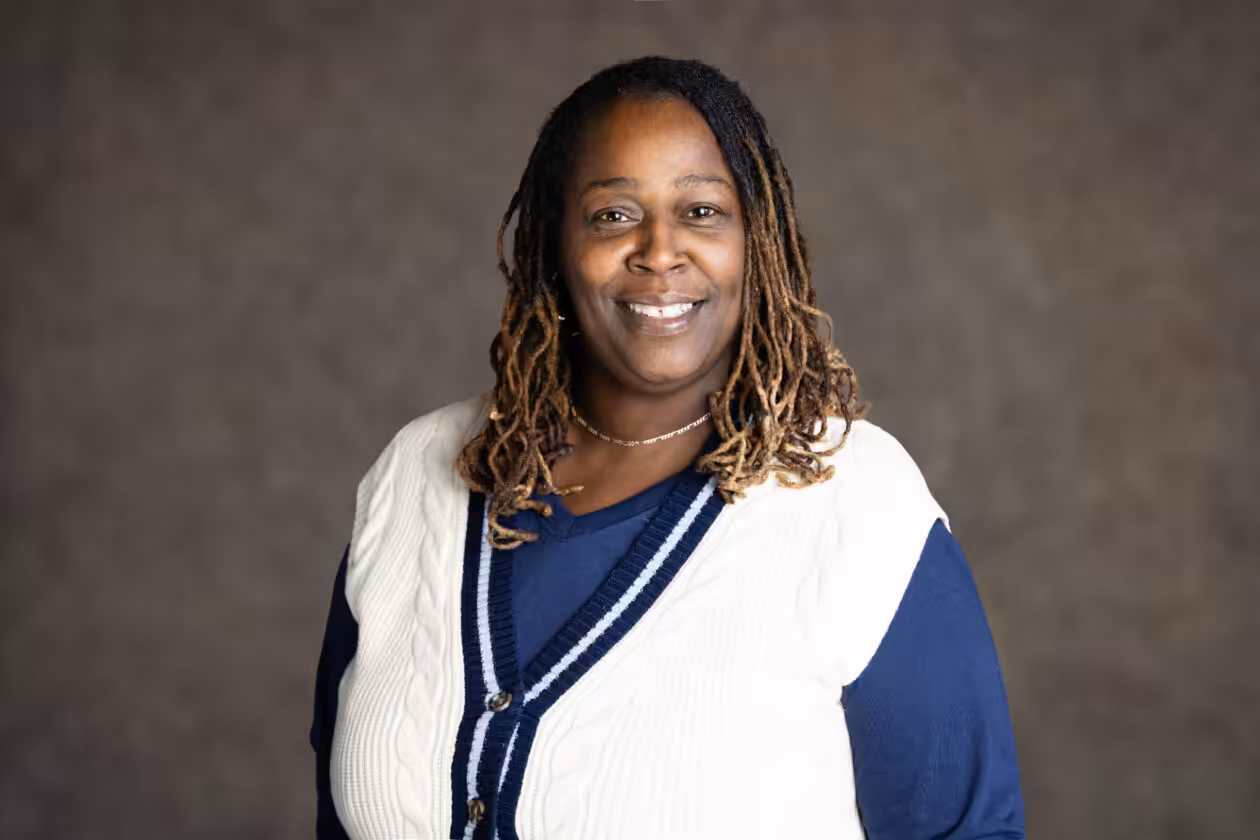 A woman with long, braided hair smiles at the camera. She is wearing a navy blue shirt and a white, sleeveless sweater with navy blue trim, standing against a plain, dark background.