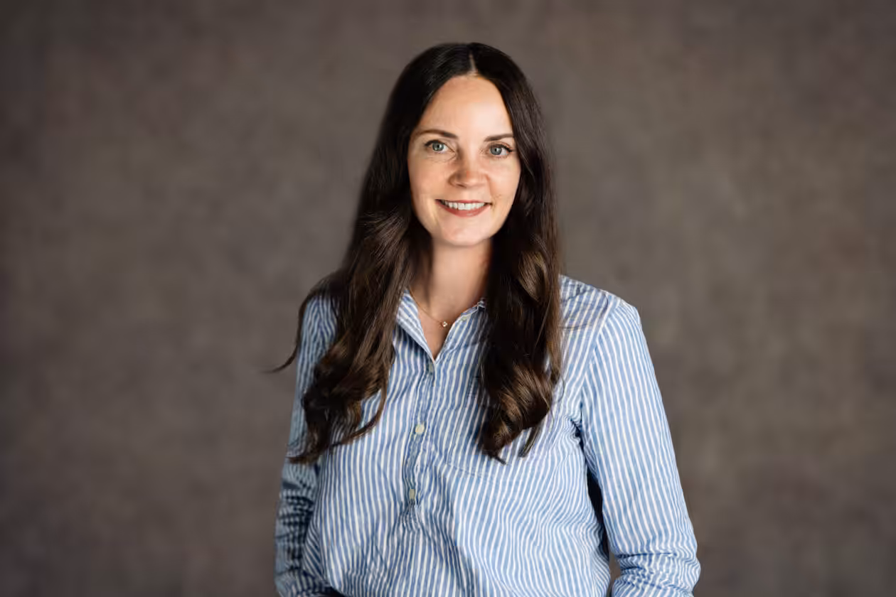A woman with long brown hair wearing a blue and white striped shirt smiles at the camera in front of a neutral, textured background.