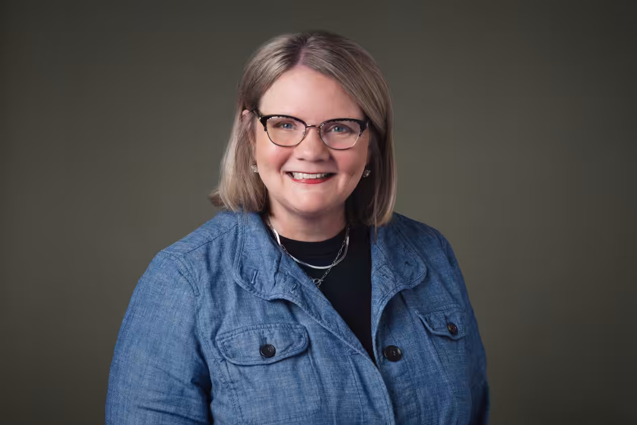 A woman with blonde hair and glasses smiles at the camera. She is wearing a blue button-up jacket over a black shirt and stands in front of a plain, dark green background.