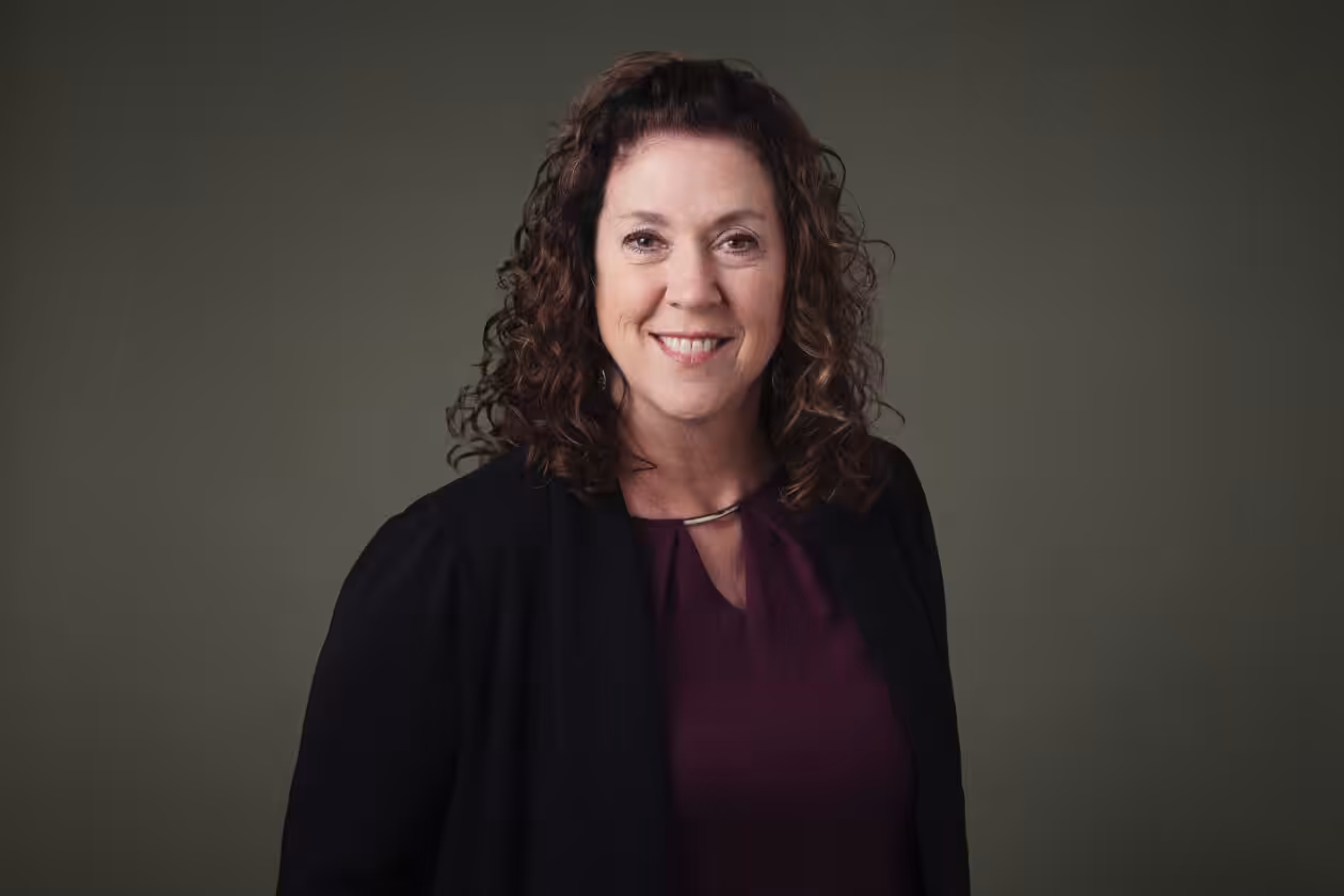 A woman with curly brown hair, wearing a dark jacket over a maroon blouse, smiles while standing in front of a plain olive green background.