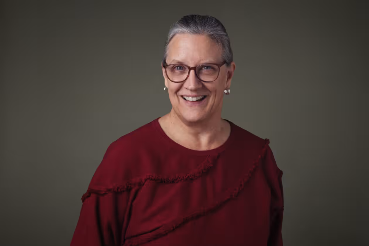 A smiling older woman with gray hair tied back, wearing glasses, pearl earrings, and a maroon top, stands against a plain olive-green background.